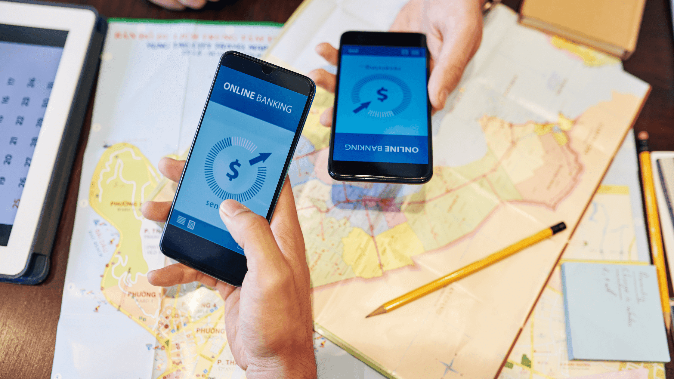 Two people holding smartphones displaying an online banking app. One phone shows a money transfer in progress. The scene includes a map, a tablet, notes, a pen, and a small dish, suggesting planning or travel activities.
